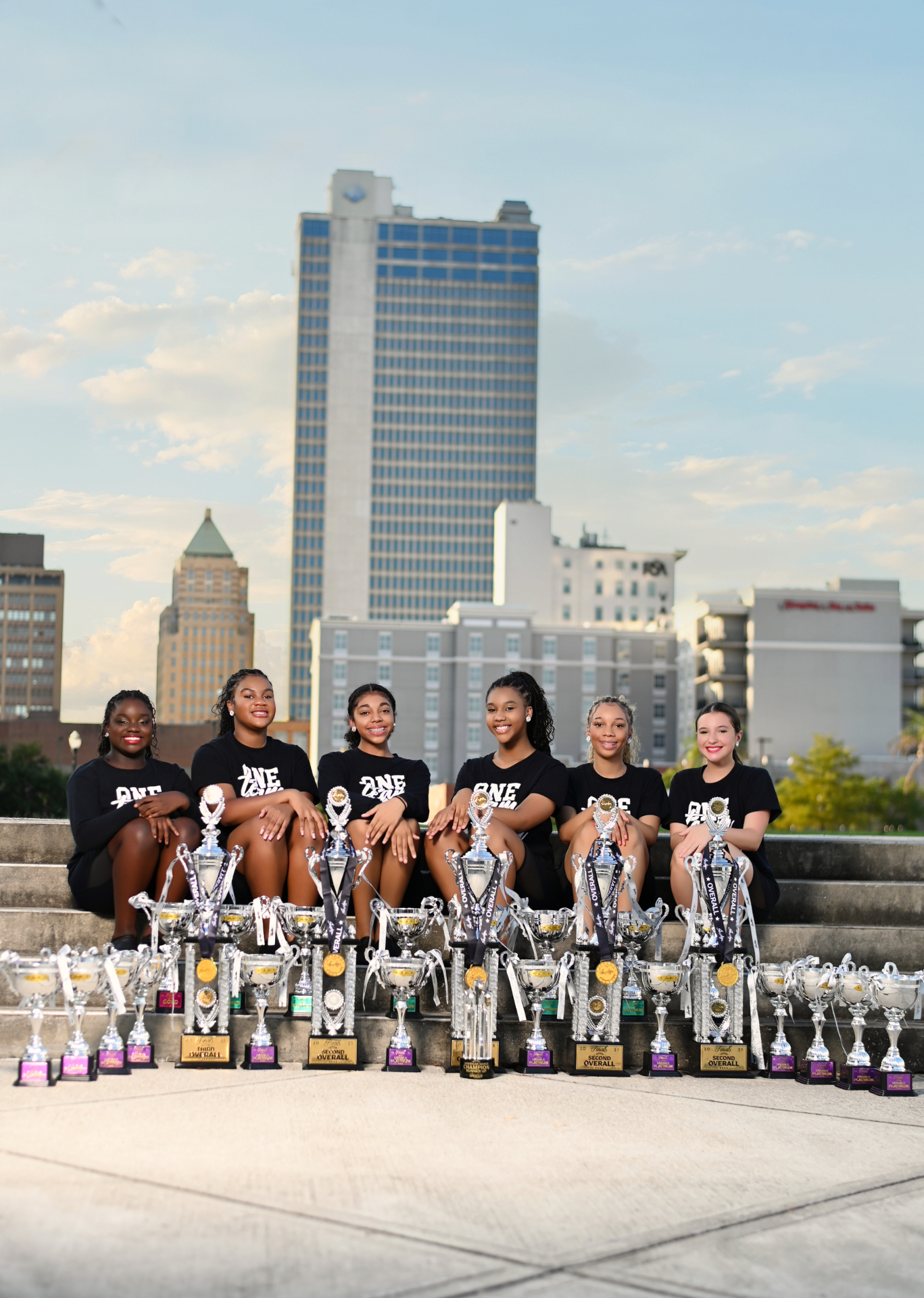 Premiere Dance Studio senior competition team seated with trophies in front of the Mobile skyline